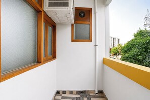 Uma varanda estreita com parede branca, janela emoldurada em madeira, uma porta pequena e chão de azulejos.