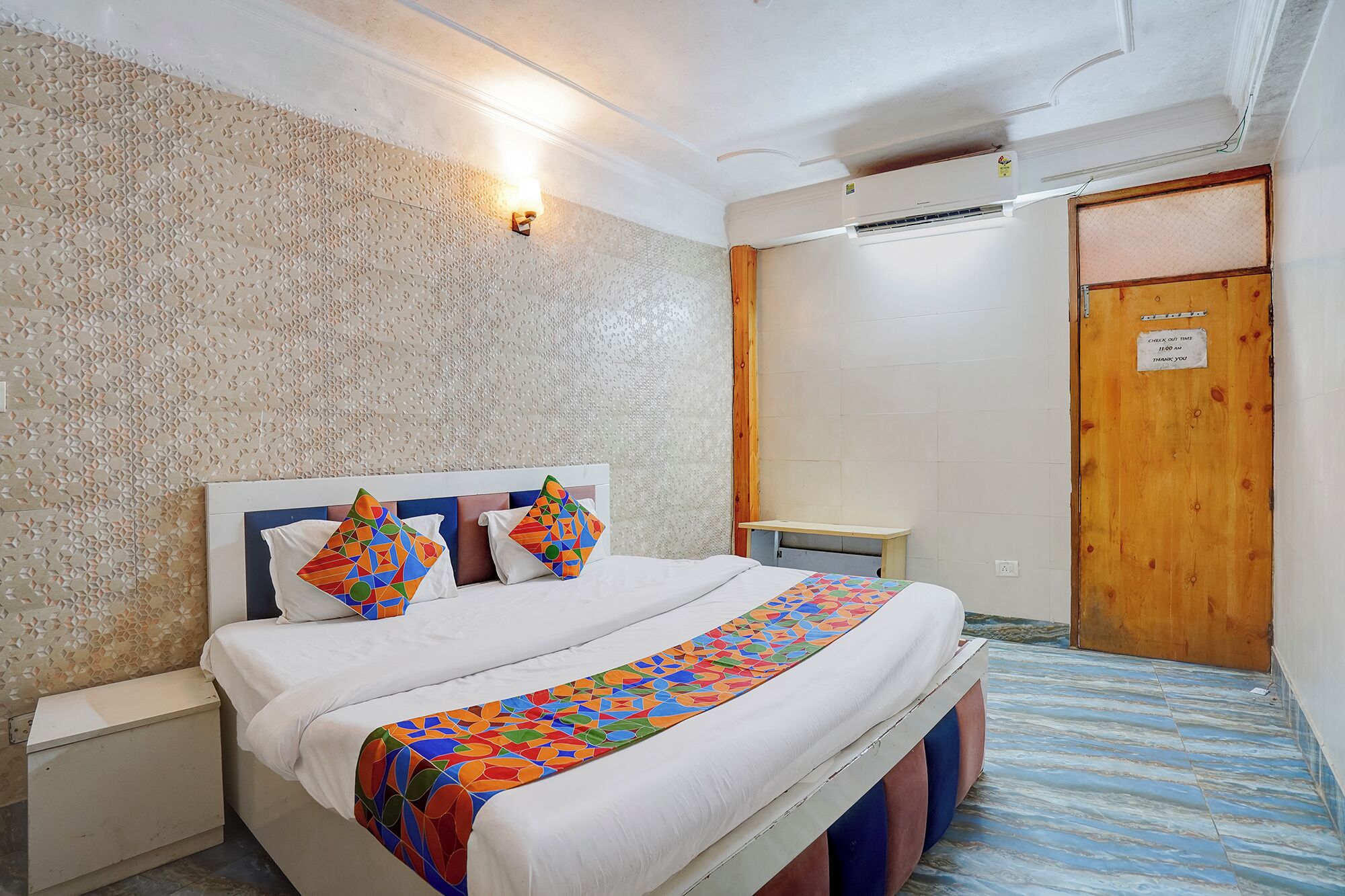 Quarto de hotel com cama, mesa de cabeceira, luminária na parede, ar condicionado e porta de madeira com uma placa.