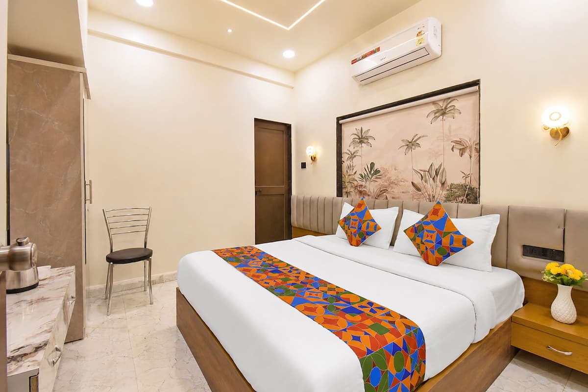 Una habitación de hotel con cama, una silla, una mesita de noche, un jarrón con flores y un equipo de aire acondicionado.