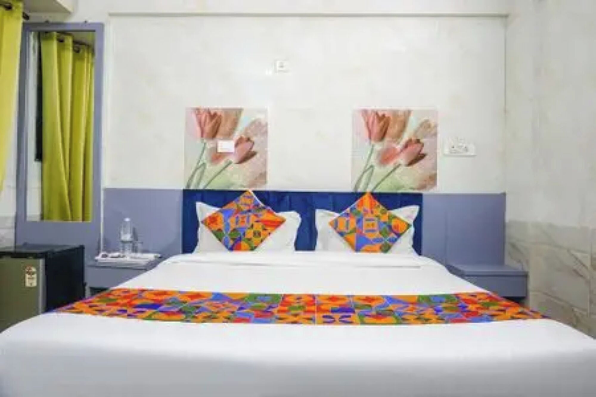 Habitación de hotel con cama, dos almohadas, mesita de noche, una botella de agua y un muro con dos cuadros.