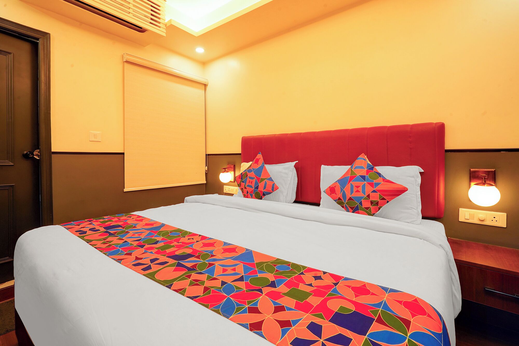 Habitación de hotel con cama, ventana con persianas, mesita de noche y lámpara de pared.