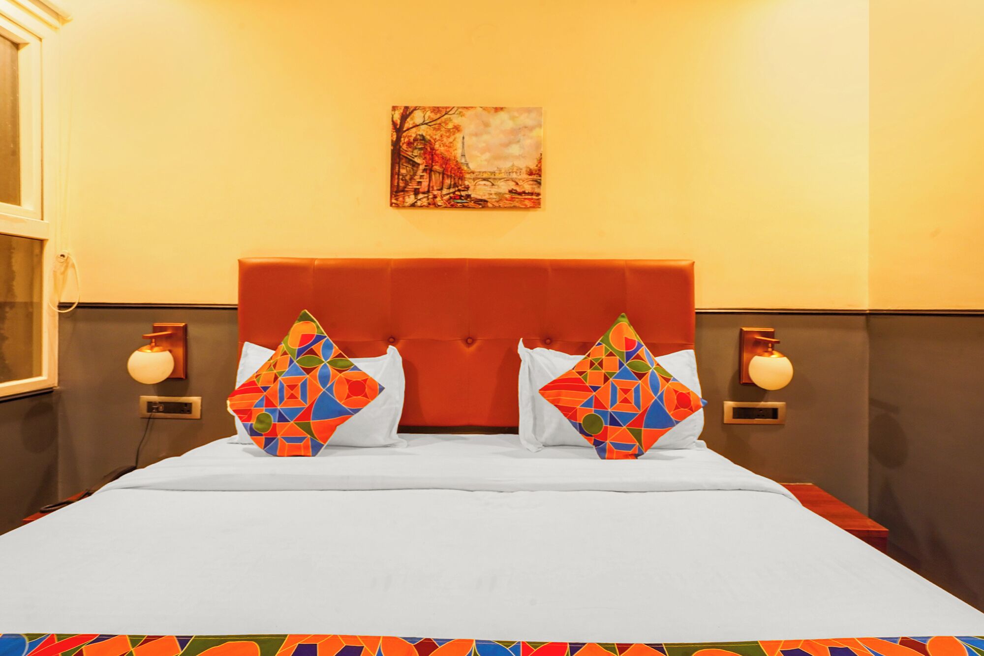 A hotel room with a large bed, two pillows with geometric patterns, a red headboard, a painting of a bridge, and wall sconces.
