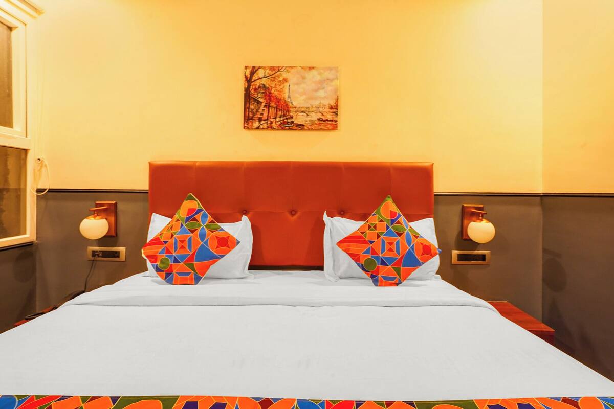 A hotel room with a large bed, two pillows with geometric patterns, a red headboard, a painting of a bridge, and wall sconces.
