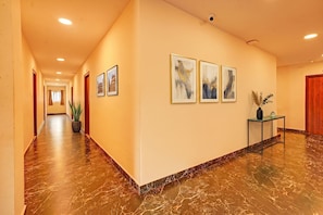 Corredor de hotel com chão de mármore, obras de arte emolduradas e um vaso de planta.