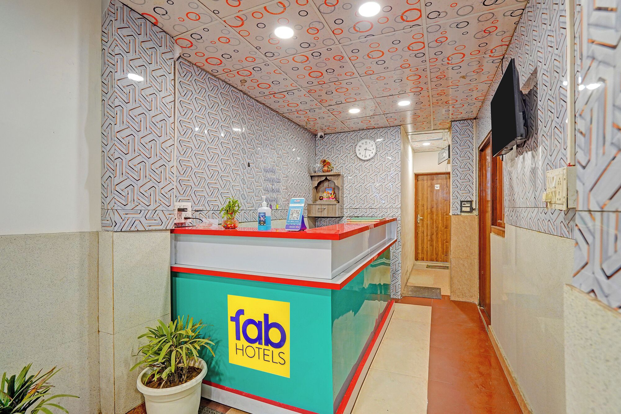 A hotel reception area with a patterned wall, a reception desk, a clock, and a potted plant.