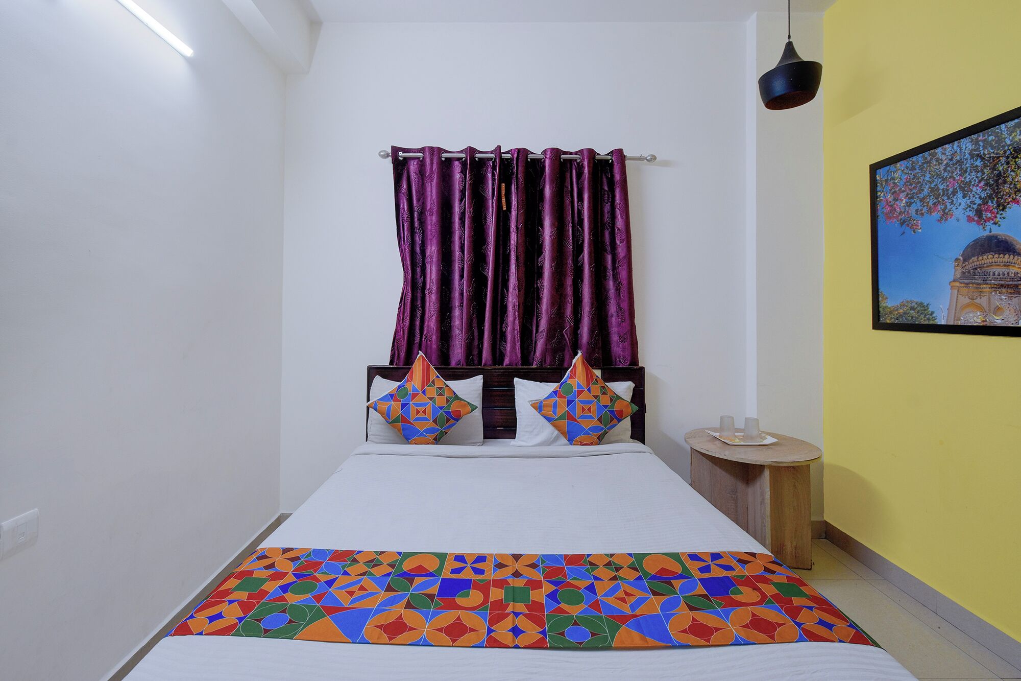 Uma cama com colcha estampada, cabeceira com almofadas geométricas, mesa de cabeceira com abajur e um quadro na parede.