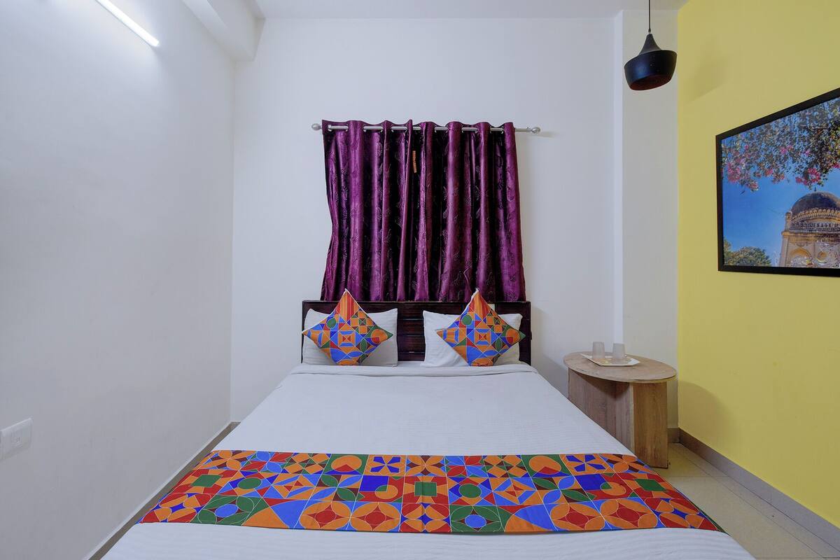 Ein Bett mit gemustertem Überwurf, ein Kopfteil mit geometrischen Kissen, ein Nachttisch mit Lampe und ein Gemälde an der Wand.