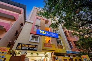 Un immeuble à plusieurs étages, sur lequel on peut voir une enseigne bien visible indiquant « Tab Hotels », une autre indiquant « Grand Broholic Luxury Rooms » et une troisième indiquant « Beautiful Stay Homes ».