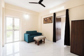 A room with a teal sofa, a small table with a patterned tablecloth, a painting on the wall, a ceiling fan, and a door with curtains. - Fabhotel Himalay Guest House (Pune)