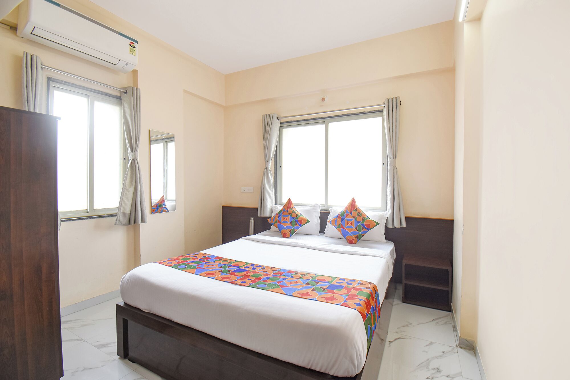 A hotel room with a bed, two windows with curtains, an air conditioning unit, and a mirror.