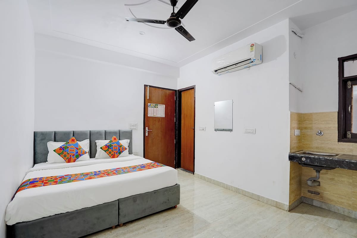 A bedroom with a bed, a ceiling fan, an air conditioning unit, a wooden door, a mirror, and a bathroom with a sink.