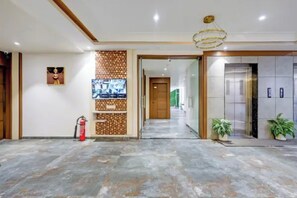 A hotel lobby with a reception desk, a fire extinguisher, a wall-mounted TV, and a hallway leading to an elevator. - Fabhotel Mh Grand (Kalol)