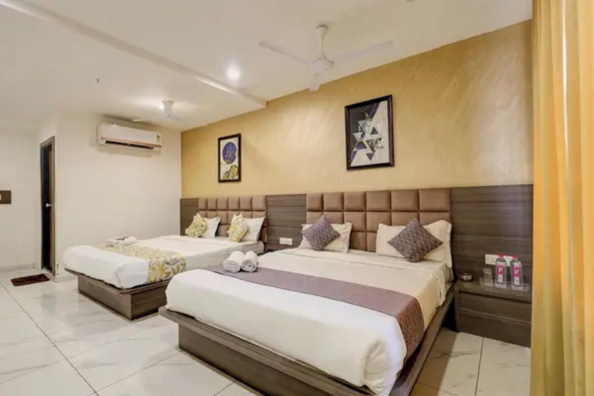 A hotel room with two beds, a ceiling fan, an air conditioning unit, and artwork on the walls.