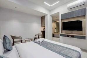 Una habitación de hotel moderna con una cama grande, un televisor montado en la pared, un pequeño rincón de descanso y un equipo de aire acondicionado.