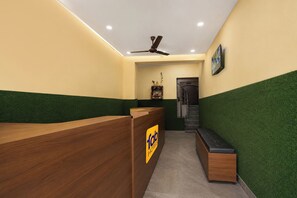 Zona de recepción de un hotel, con un mostrador de madera, un banco de asientos, un ventilador de techo y una escalera que sube al piso superior.