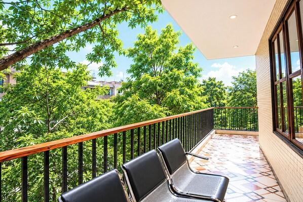 A balcony with two black chairs, a wooden railing, and a view of green trees and buildings.