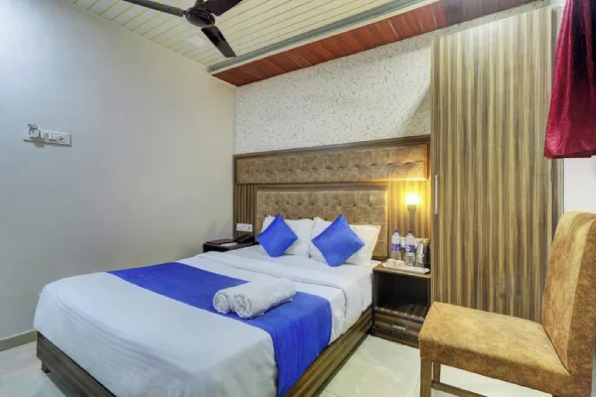 Una habitación de hotel con cama, una silla, una mesita de noche y un ventilador de techo.