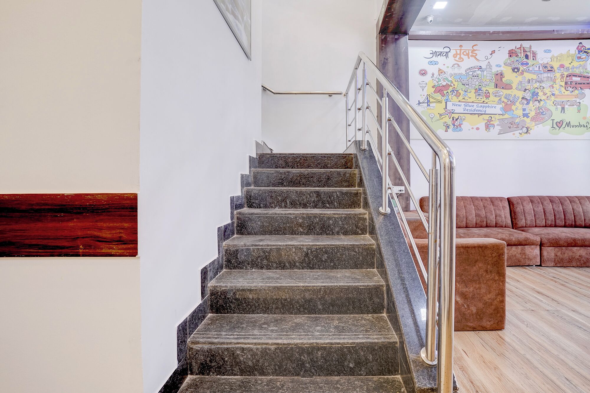 A staircase with metal railings leading to a higher floor, a brown sofa in the foreground, and a colorful wall art depicting Mumbai.