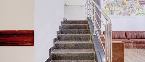 A staircase with metal railings leading to a higher floor, a brown sofa in the foreground, and a colorful wall art depicting Mumbai.