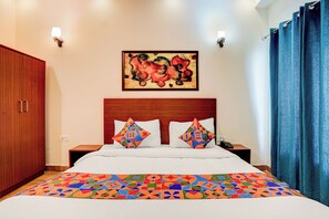 Una habitación de hotel con un armario de madera, una cama con colcha de diseño geométrico y colores vivos, dos almohadas con motivos geométricos, un cuadro abstracto en la pared y una cortina azul.