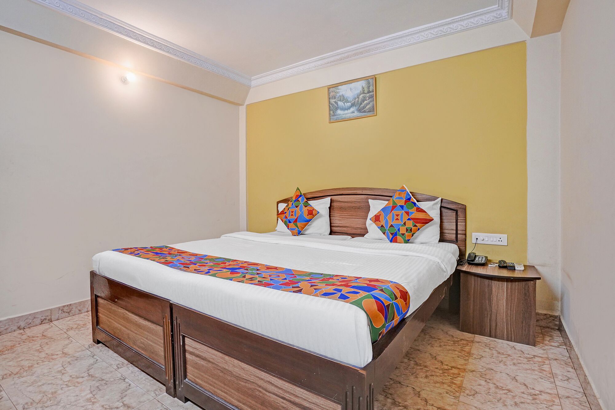 A hotel room with a bed, a bedside table, a nightstand, and a painting on the wall.