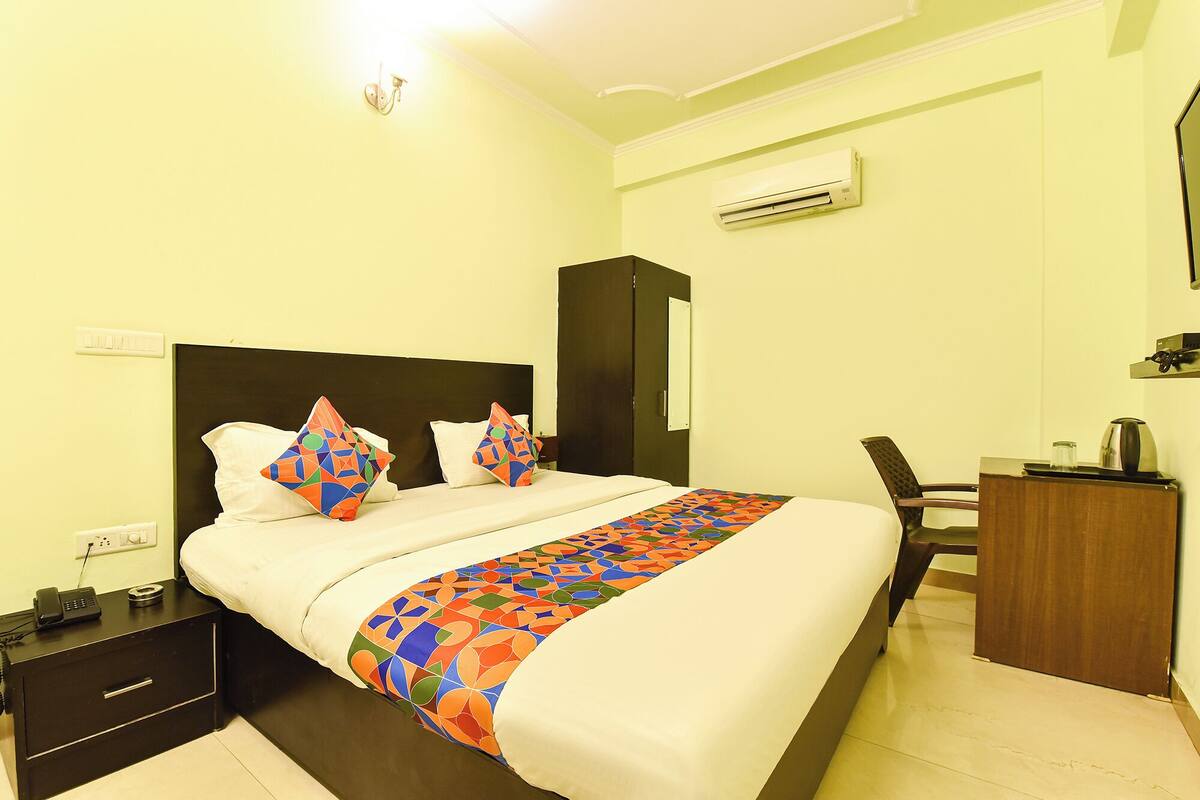 A hotel room with a bed, bedside table, nightstand, desk, chair, and an air conditioning unit.