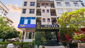 A multi-story hotel with a prominent 'HOTEL RAGHUKUL' sign, a visible entrance with steps, and a surrounding garden with potted plants.