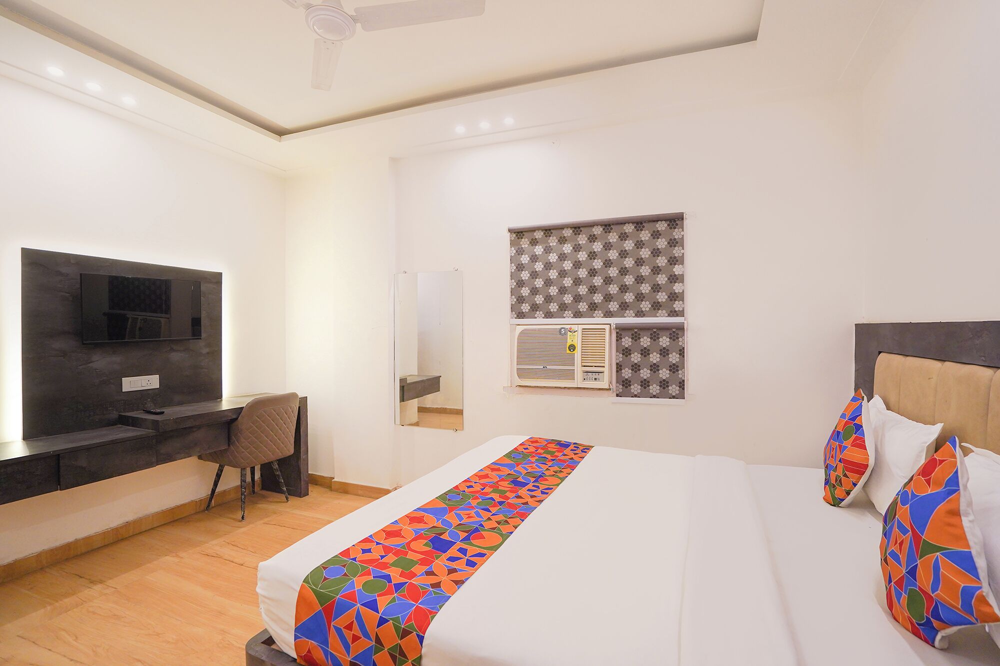 A hotel room with a bed, a desk, a TV, and a window with patterned curtains.