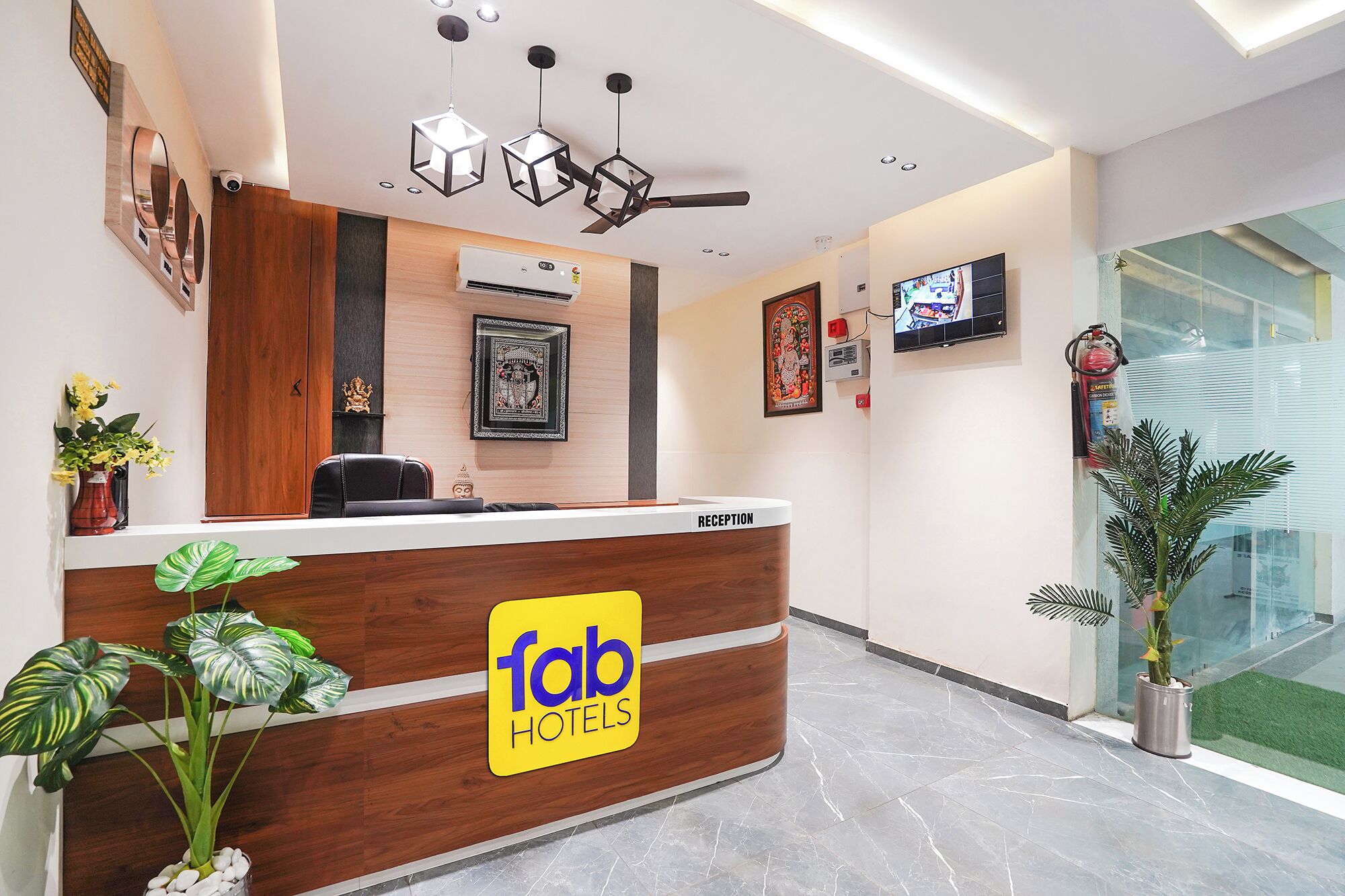 A hotel reception desk with a wooden finish, a reception area, a ceiling fan, a mounted television, and a large window.
