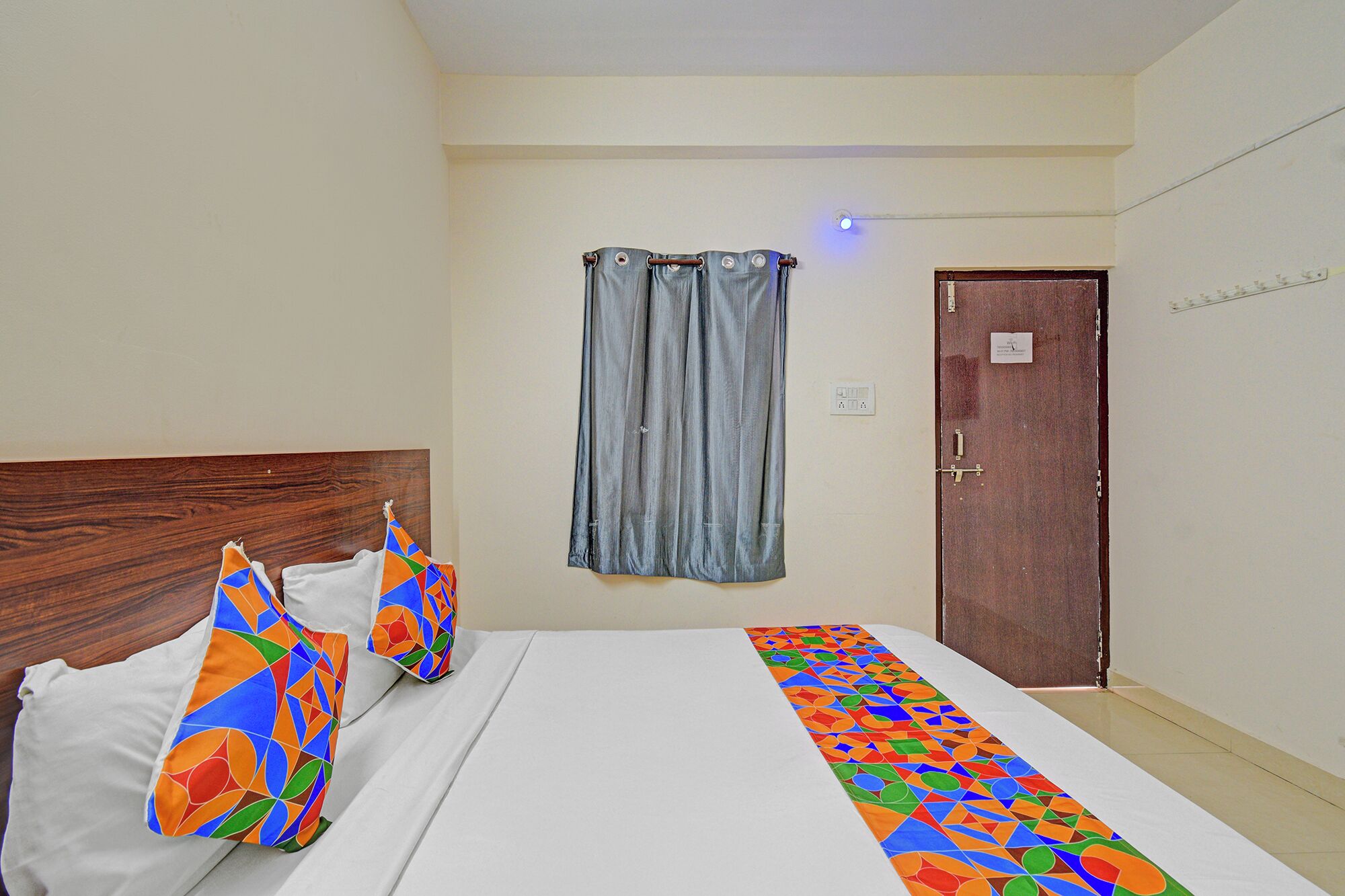 A hotel room with a wooden headboard, a bed with white bedding, colorful pillows, a blue curtain, and a wooden door.