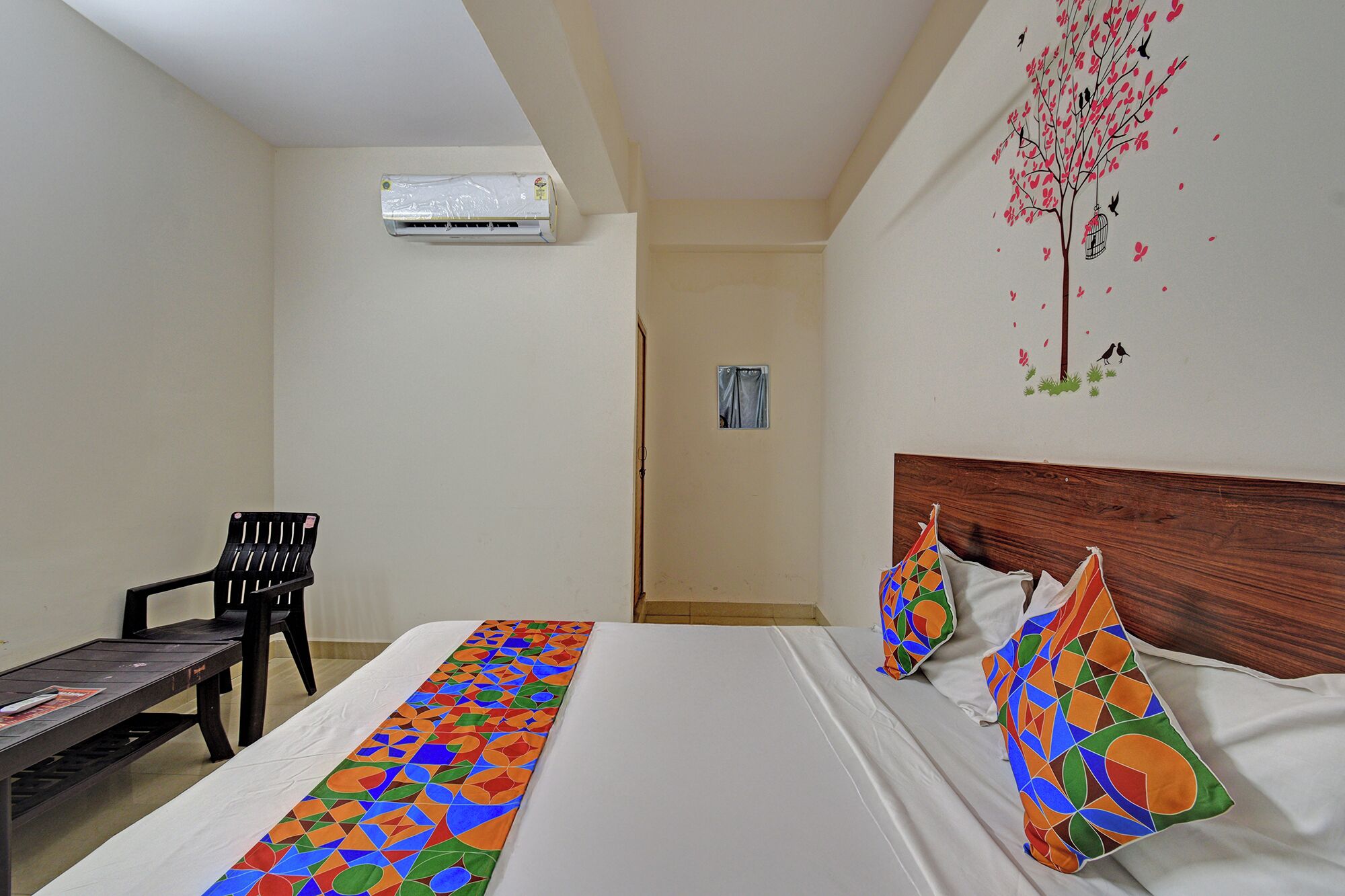 A hotel room with a bed, a chair, a table, an air conditioning unit, and a wall with a tree and birds mural.