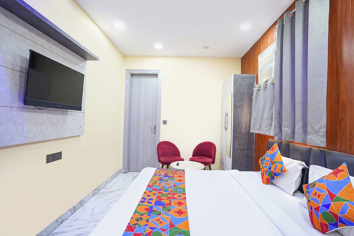 A hotel room with a bed, a TV mounted on the wall, two red chairs, a small table, and a window with curtains.