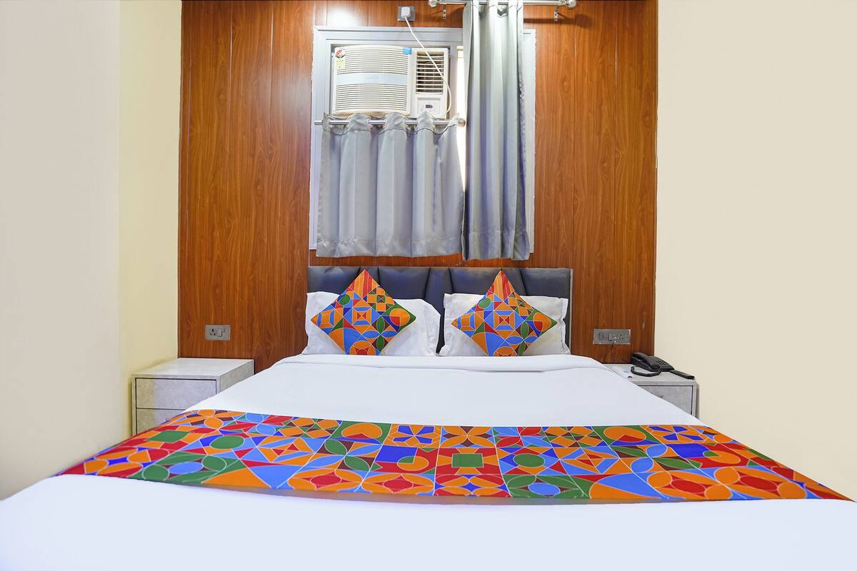 A hotel room with a bed, two pillows, a bedside table, a phone, and an air conditioning unit.
