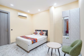 A hotel room with a bed, a round table, a green chair, a wardrobe, and an air conditioning unit. - Fabhotel Shelter Inn (New Delhi)