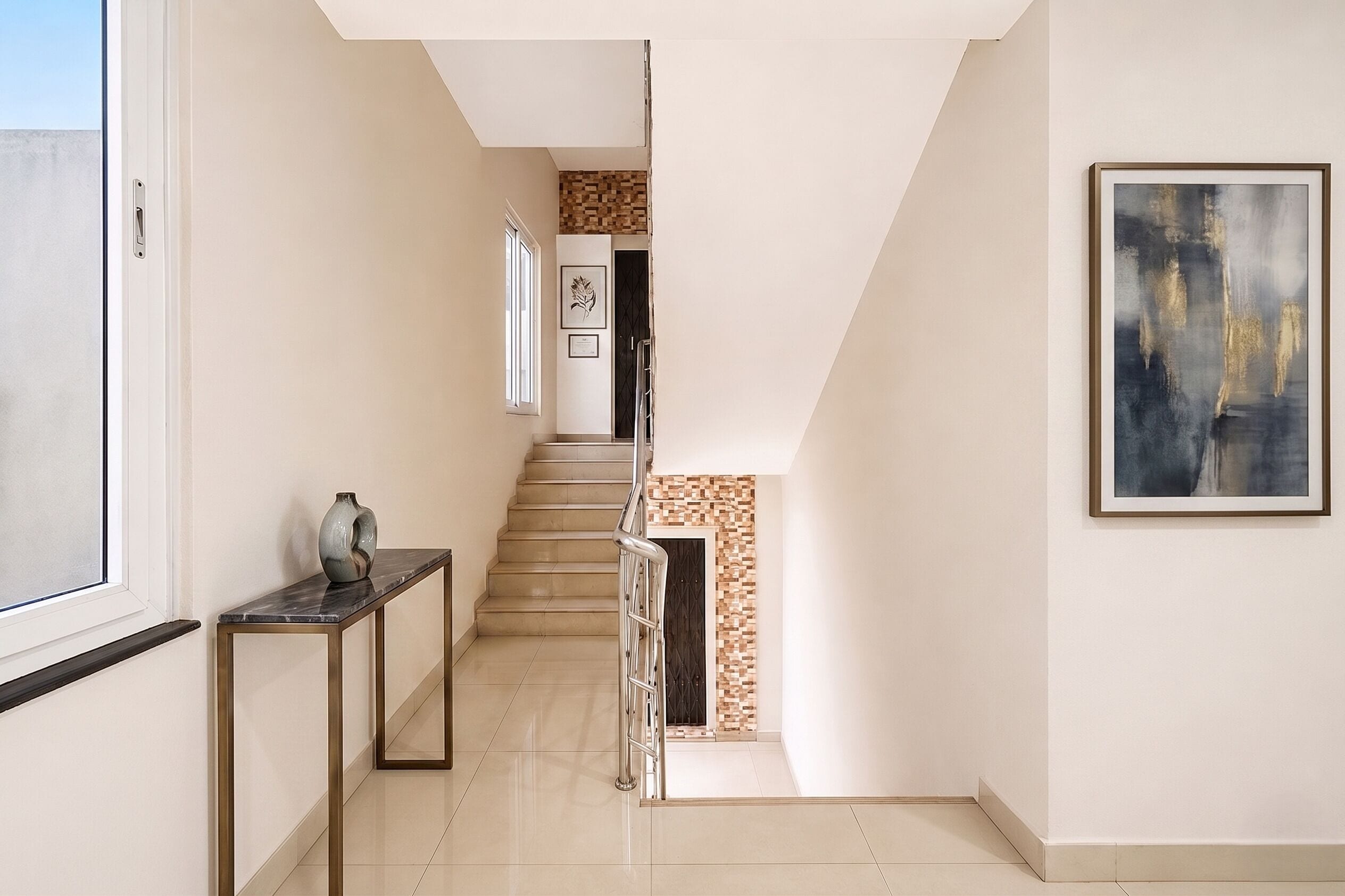 Un couloir avec un escalier, une petite table sur laquelle est posé un vase, et un tableau encadré accroché au mur.