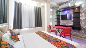 Una habitación de hotel con cama, televisor montado en la pared, una mesa de comedor con sillas y una ventana con cortinas.