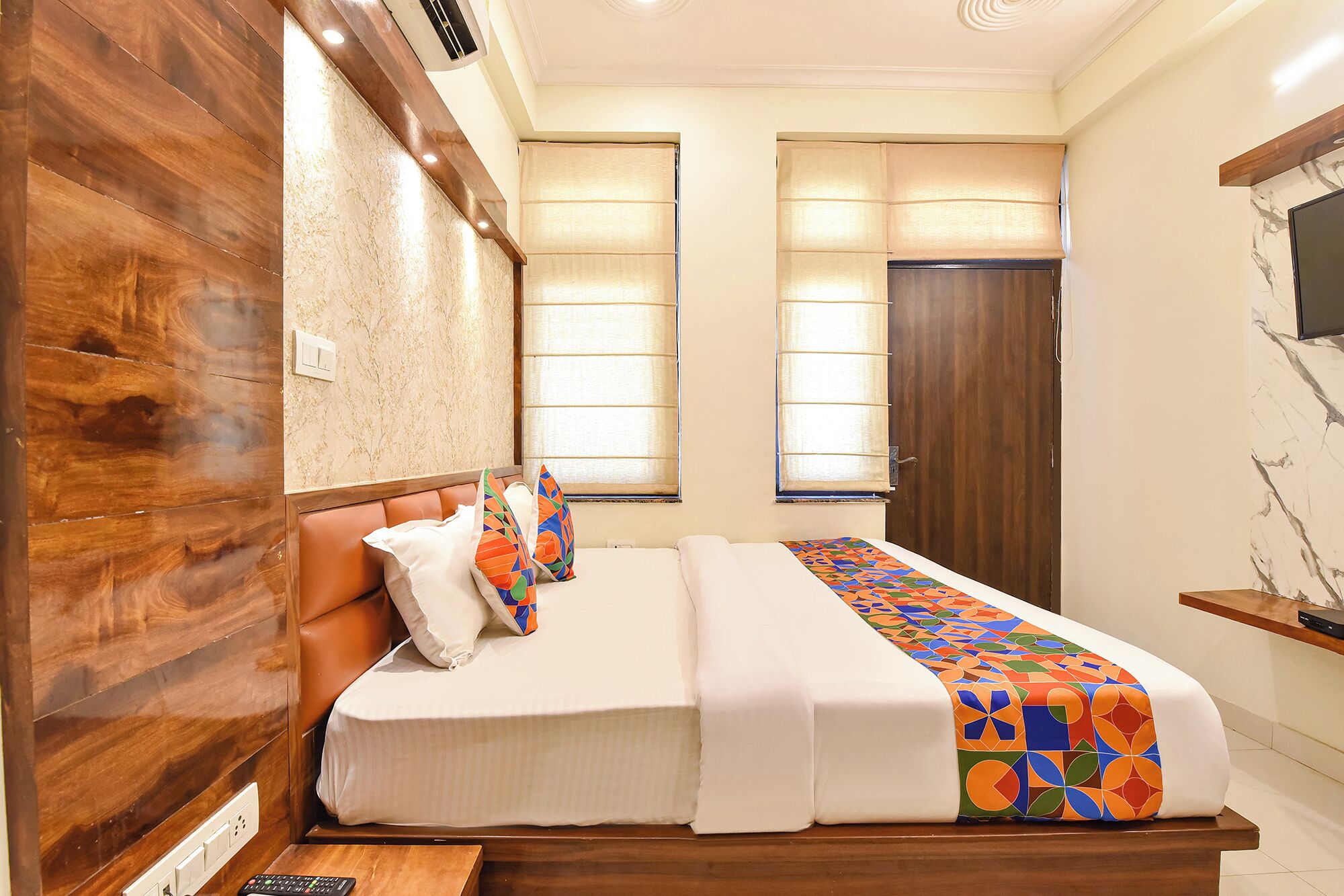 A bedroom with a wooden headboard, a bed with white bedding, two windows with curtains, and a TV mounted on the wall.