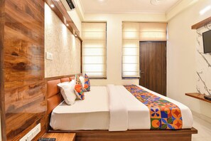 A bedroom with a wooden headboard, a bed with white bedding, two windows with curtains, and a TV mounted on the wall.