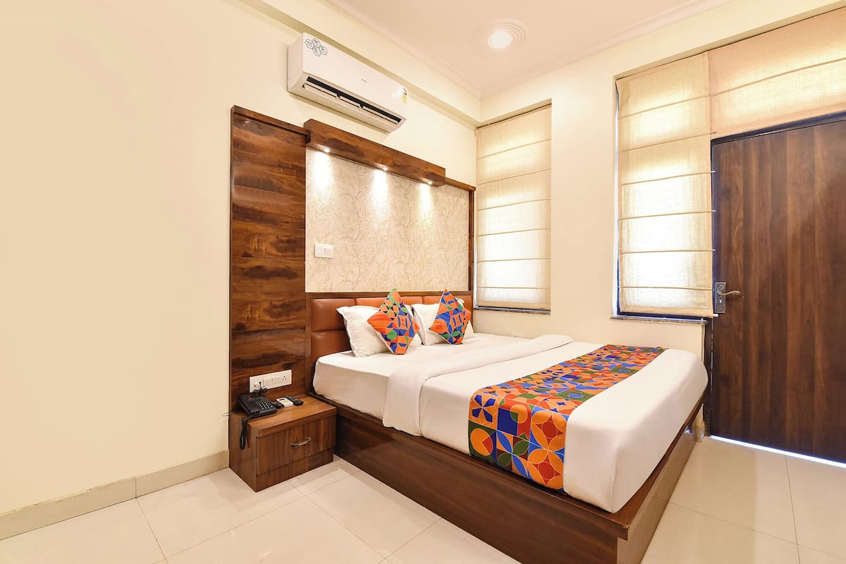 A hotel room with a bed, bedside table, air conditioning unit, and a window with blinds.