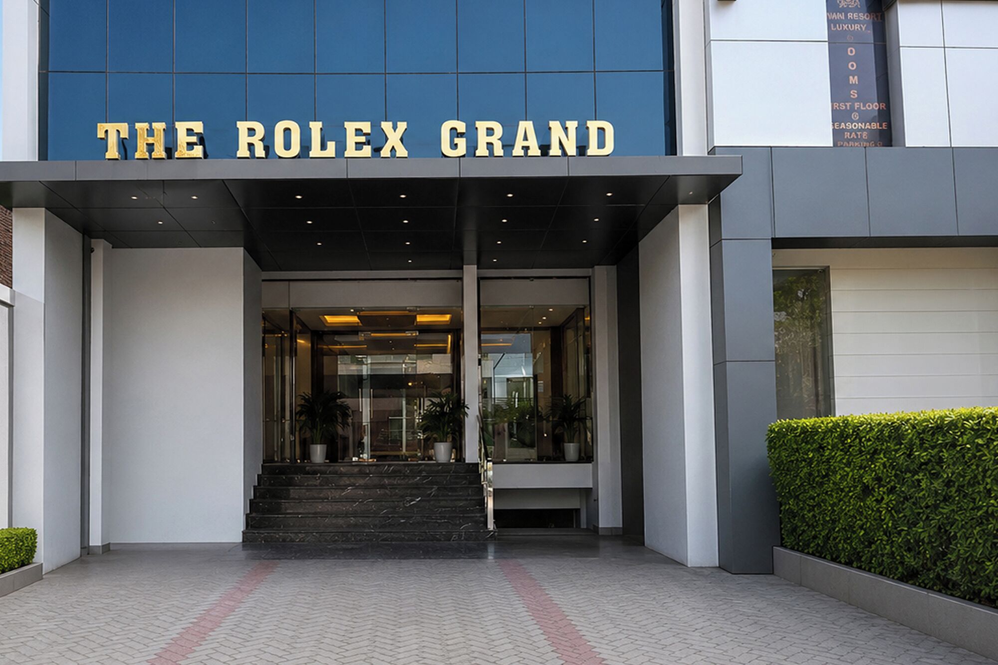 A entrada do Hotel Rolex Grand, com fachada moderna, portas de vidro e uma escadaria que leva à entrada.