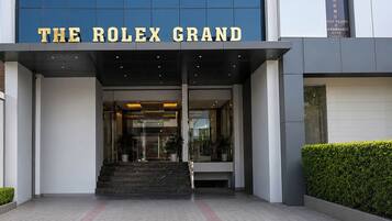 A entrada do Hotel Rolex Grand, com fachada moderna, portas de vidro e uma escadaria que leva à entrada.