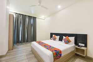 A hotel room with a bed, a nightstand, a ceiling fan, and a view of the outside through a window with curtains. - Fabhotel The Rolex Grand (Faridabad)