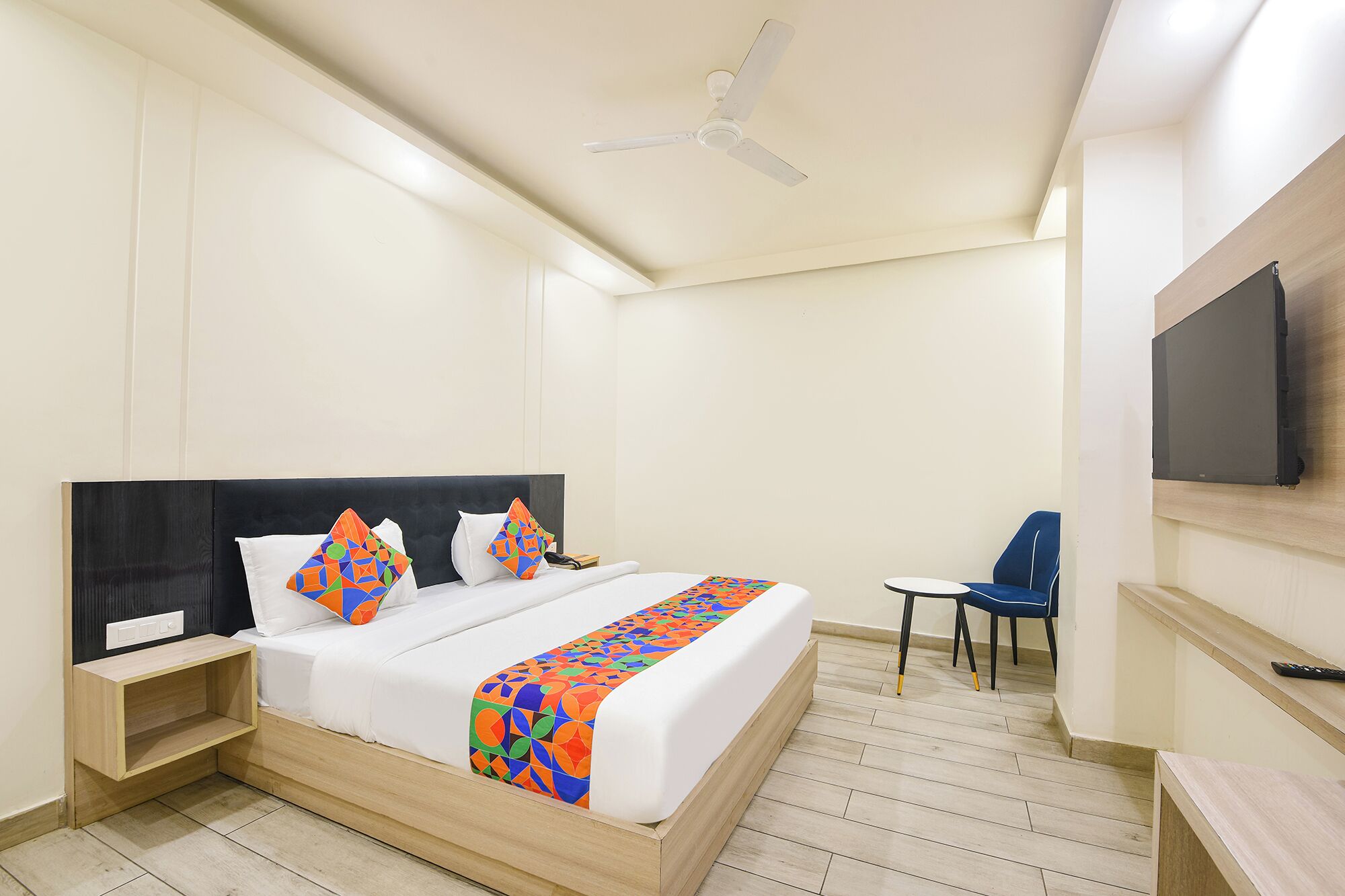 A hotel room with a bed, a bedside table, a chair, a TV mounted on the wall, and a ceiling fan.