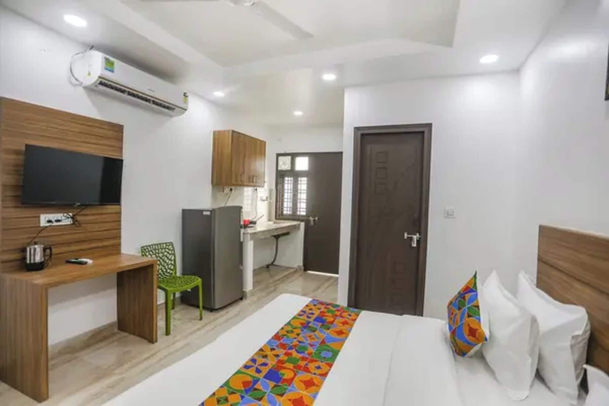 A hotel room with a bed, a desk with a computer, a chair, a TV mounted on the wall, and an air conditioning unit.