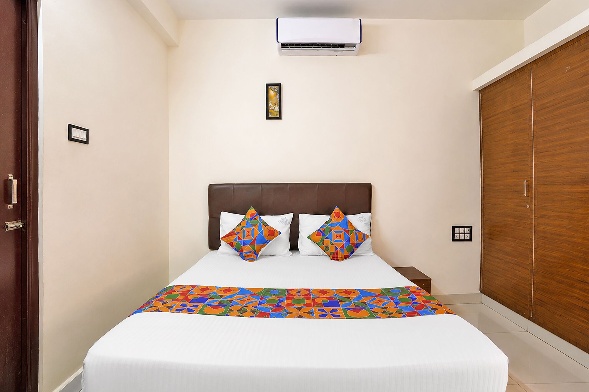 A bedroom with a bed, pillows, an air conditioner, a wooden wardrobe, and a framed picture on the wall.