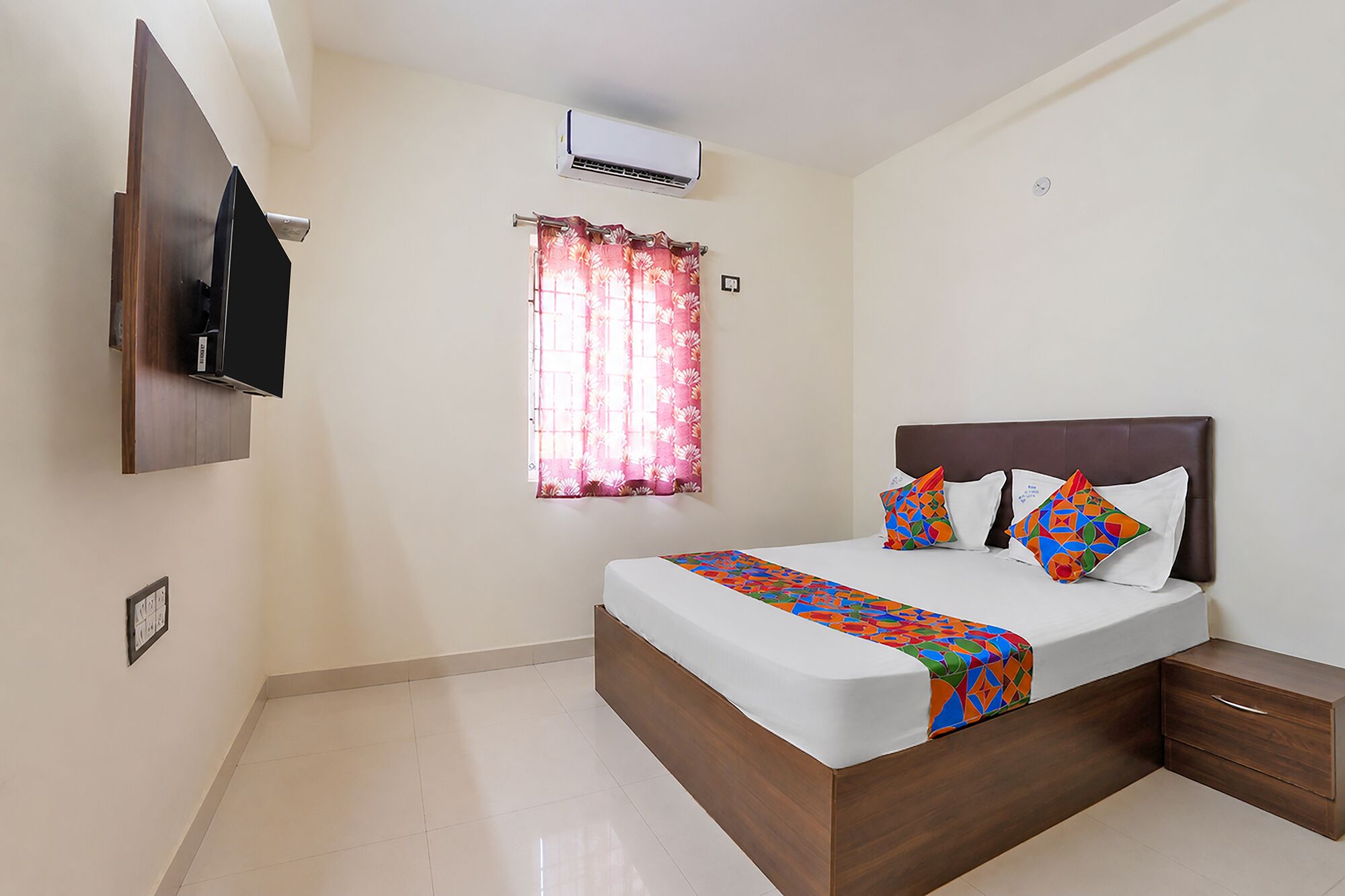 A bedroom with a bed, a TV mounted on the wall, an air conditioner, and a window with curtains.