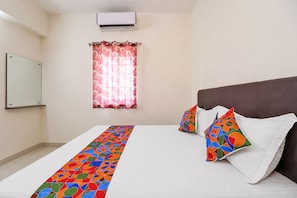 A hotel room with a bed, a window with a curtain, an air conditioning unit, and a wall-mounted mirror.