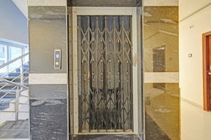 Porta de elevador de metal com um padrão geométrico, escadaria à esquerda e parede lisa à direita.