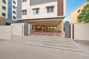 Entrada de um edifício com um caminho de azulejos que leva a uma área coberta com portas de vidro.