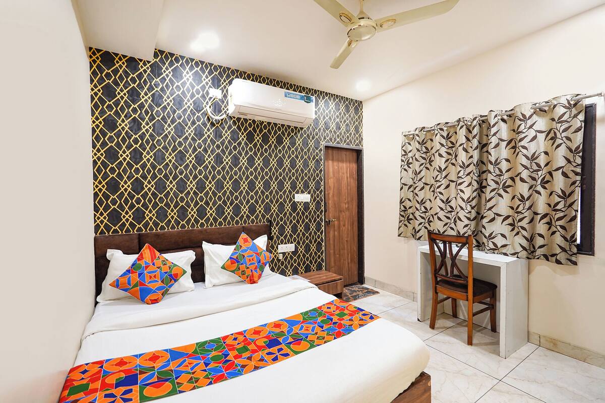 A hotel room with a bed, a desk, a chair, an air conditioning unit, and a window with curtains.